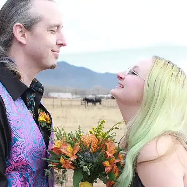 A newly married couple smiles at each other with a couple horses in the background